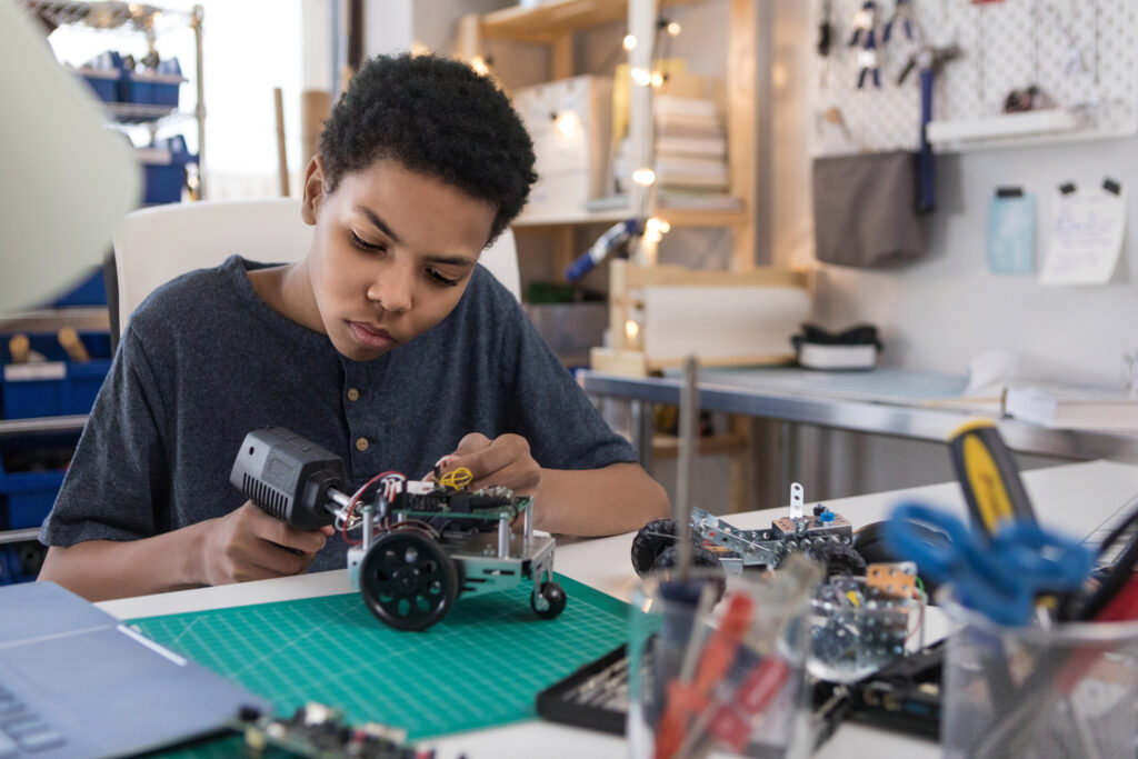 A serious teen boy uses a soldering gun to connect wires as he builds a robot at home.